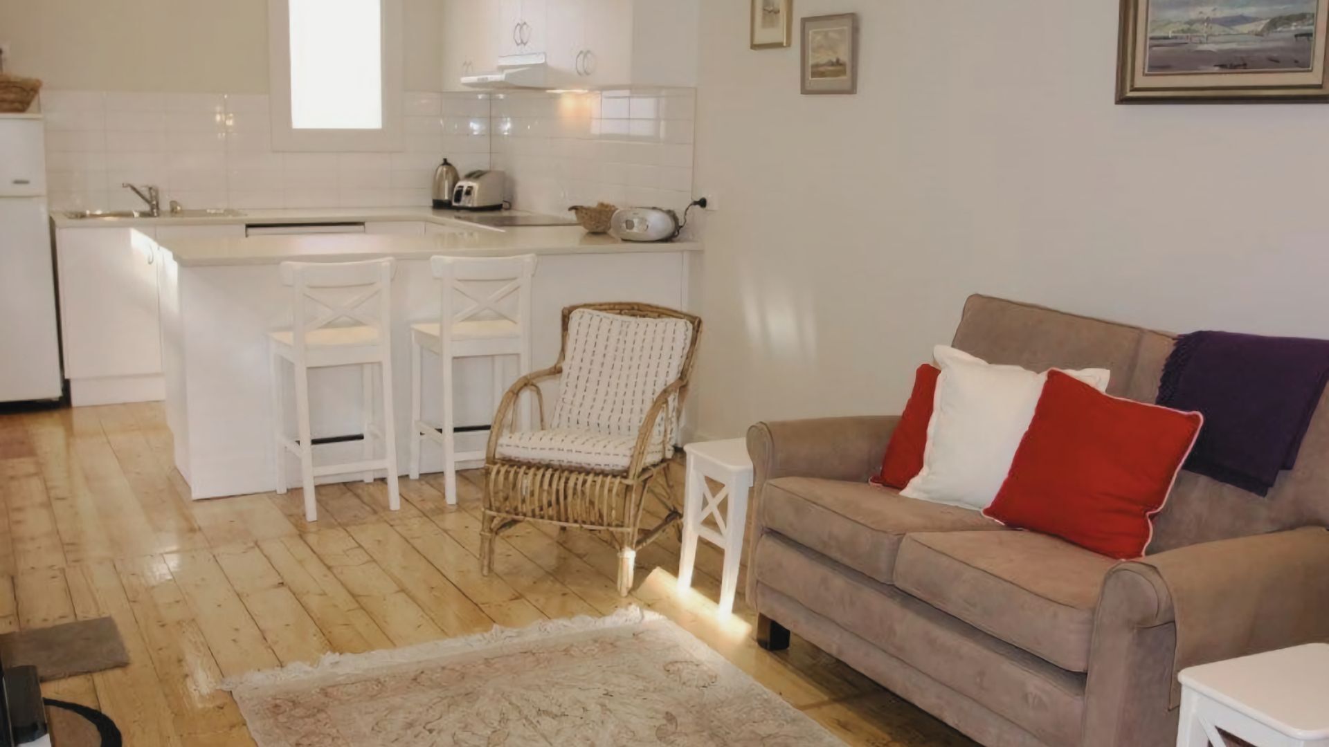 En kompakt opholdsstue med tekøkken, en fletstol, en sofa med røde og hvide puder og en lille måtte.