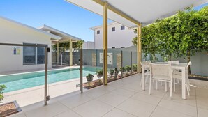 A modern outdoor pool area with a glass enclosure, a white table and chairs, and a small garden with trees and shrubs. - Splash House at Kingscliff - Pet Friendly with Pool (KINGSCLIFF)