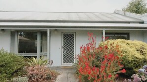 A single-story house with a front porch, a white door with a metal grille, and a variety of flowering plants in the front yard.