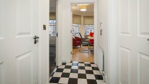 A narrow hallway with a checkered floor leading to a living area with a red chair and a table. - Haven Apartments at Eglantine Avenue (Belfast)