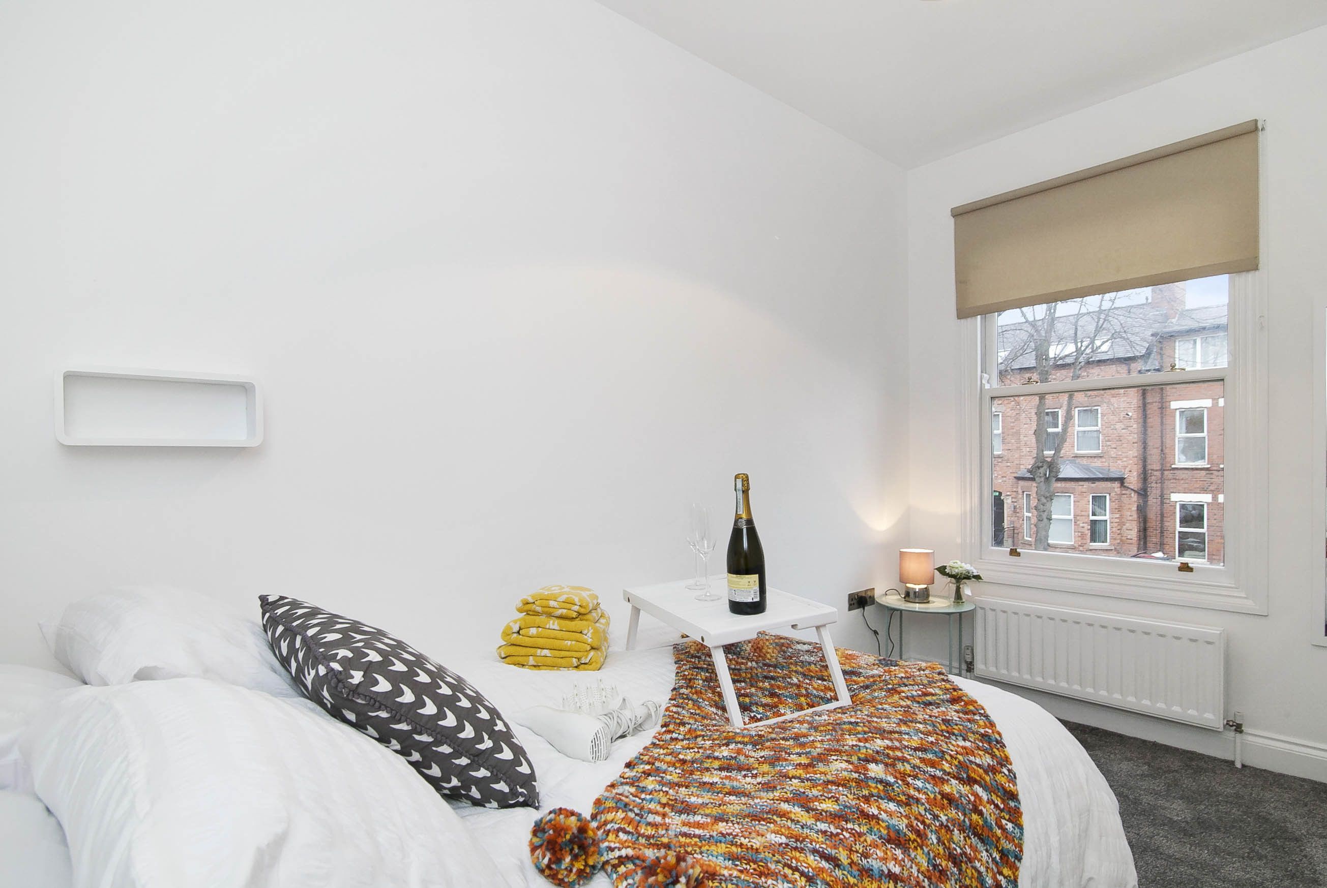 A bedroom with a bed, a bedside table with a bottle and glass, a window with a view of buildings, and a radiator.