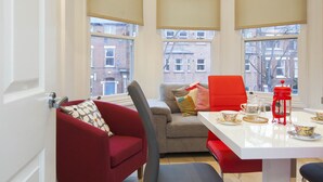 A cozy corner room with a red sofa, a grey armchair, and a white table with a red lantern. Large windows offer a view of the city. - Haven Apartments at Eglantine Avenue (Belfast)