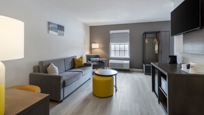 Een moderne woonkamer met een grijze bank, een geel poefje, een flatscreen televisie en een keuken met een donker aanrecht.