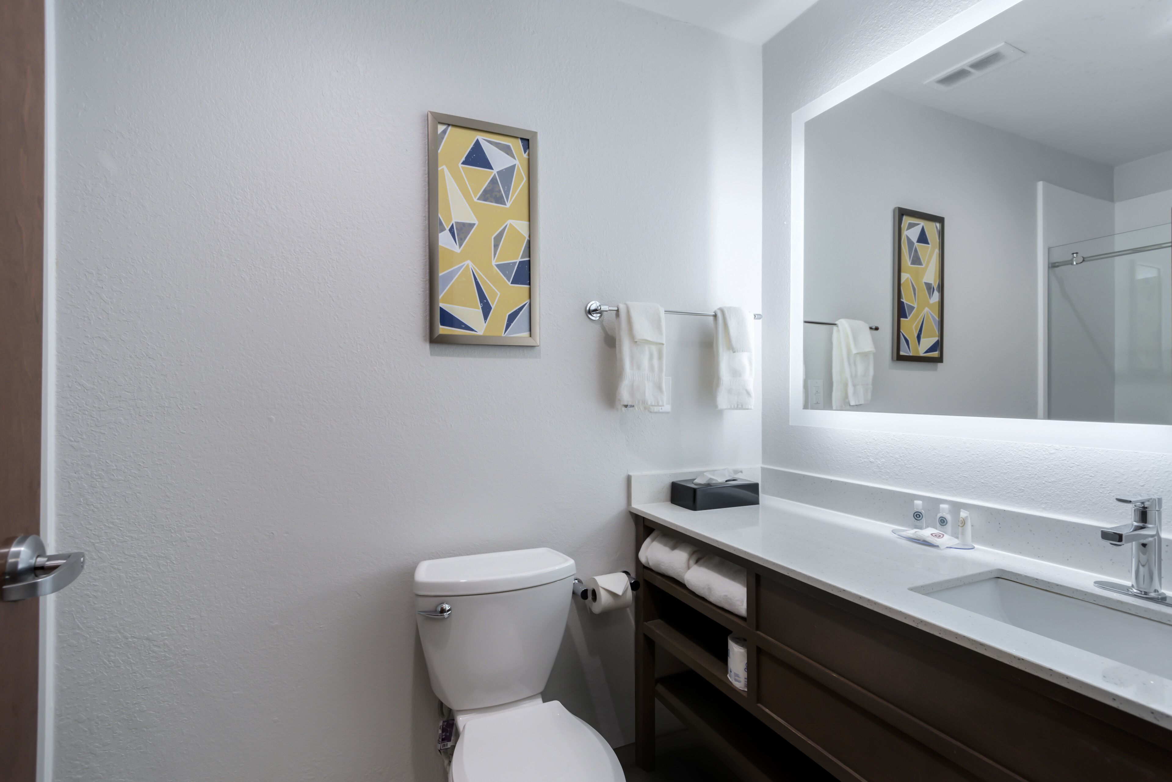 Un baño con inodoro, lavamanos y espejo. Hay toallas colgadas en la pared y un cuadro enmarcado sobre el espejo.