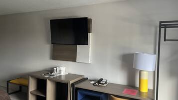 A hotel room with a bed, a desk with a telephone and a lamp, a TV mounted on the wall, and a small table with a cup and a phone.