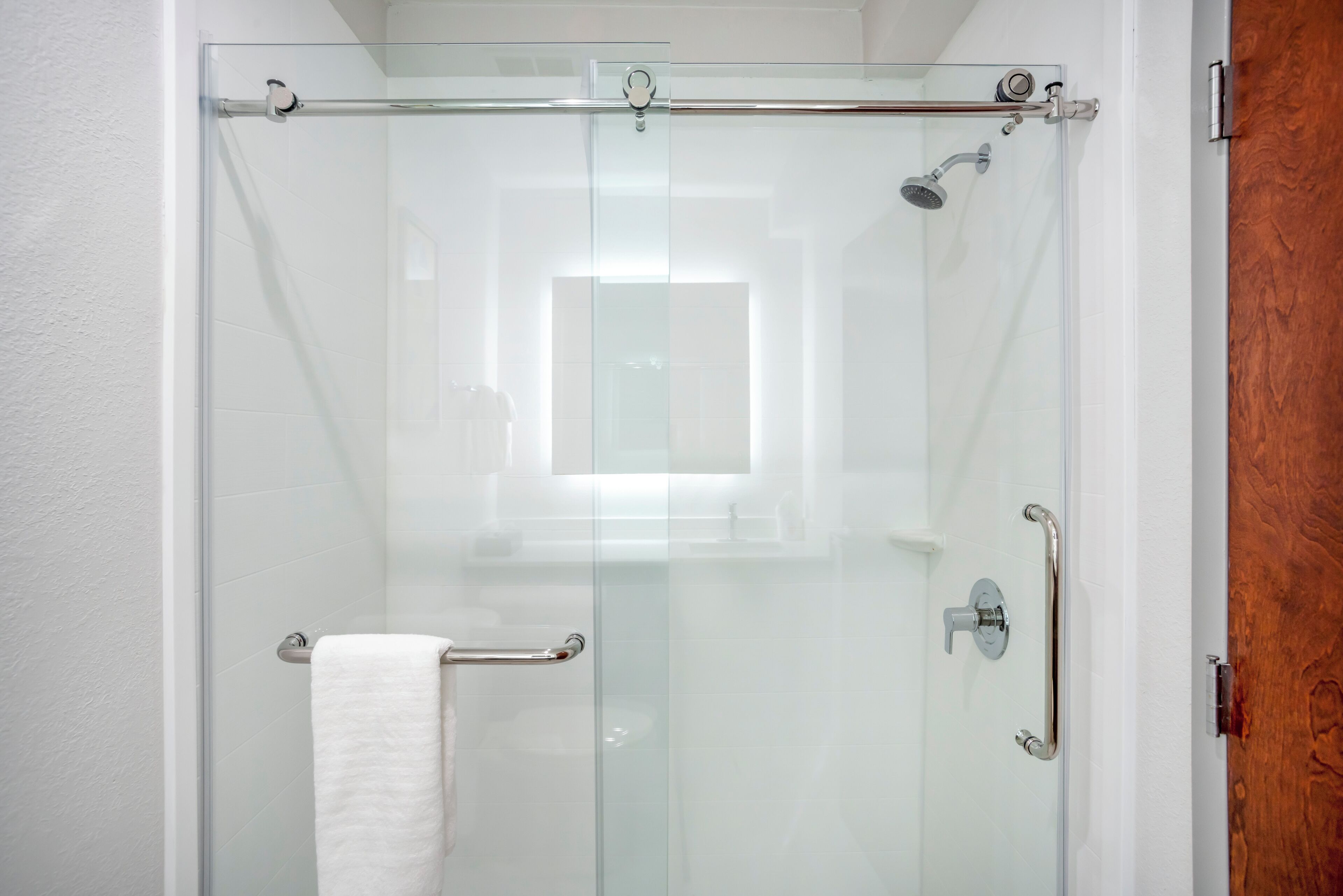 A glass-enclosed shower with a white towel on the door handle, a glass door, and a wooden door frame.