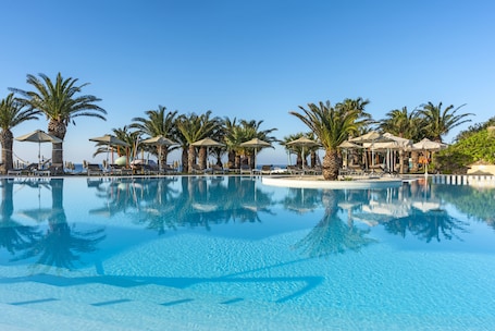 A clear blue pool with palm trees and umbrellas, surrounded by lounge chairs and a clear sky.