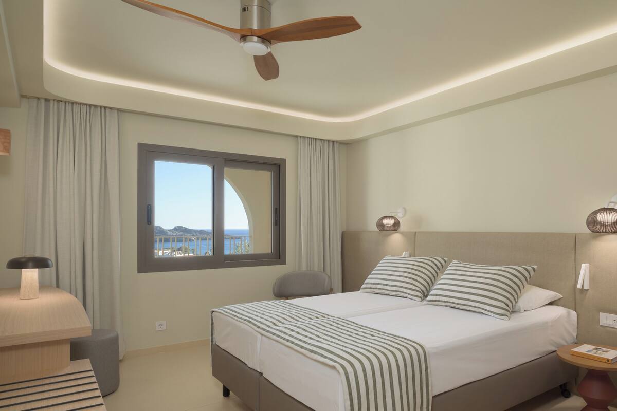 Habitación de hotel con cama, escritorio, silla, ventilador de techo, ventana con cortinas y vista al mar.