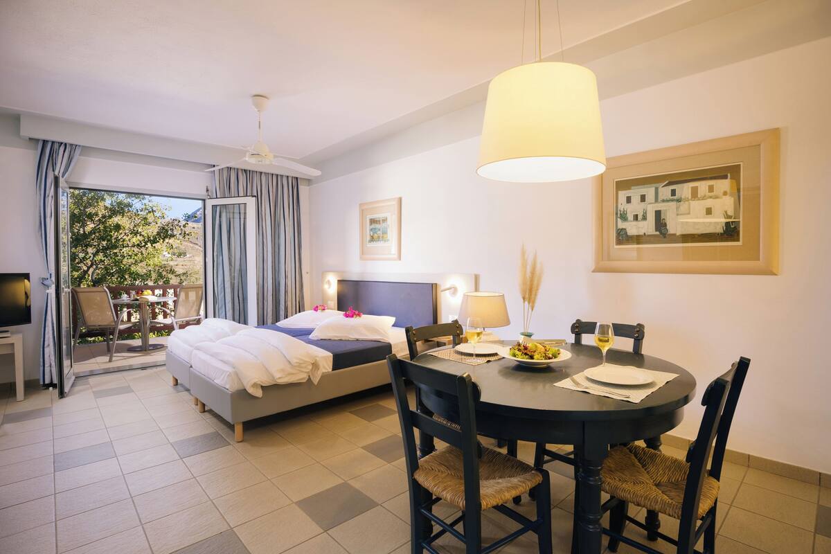Habitación de hotel con cama, una mesa de comedor con sillas, televisor y balcón con vistas a árboles.