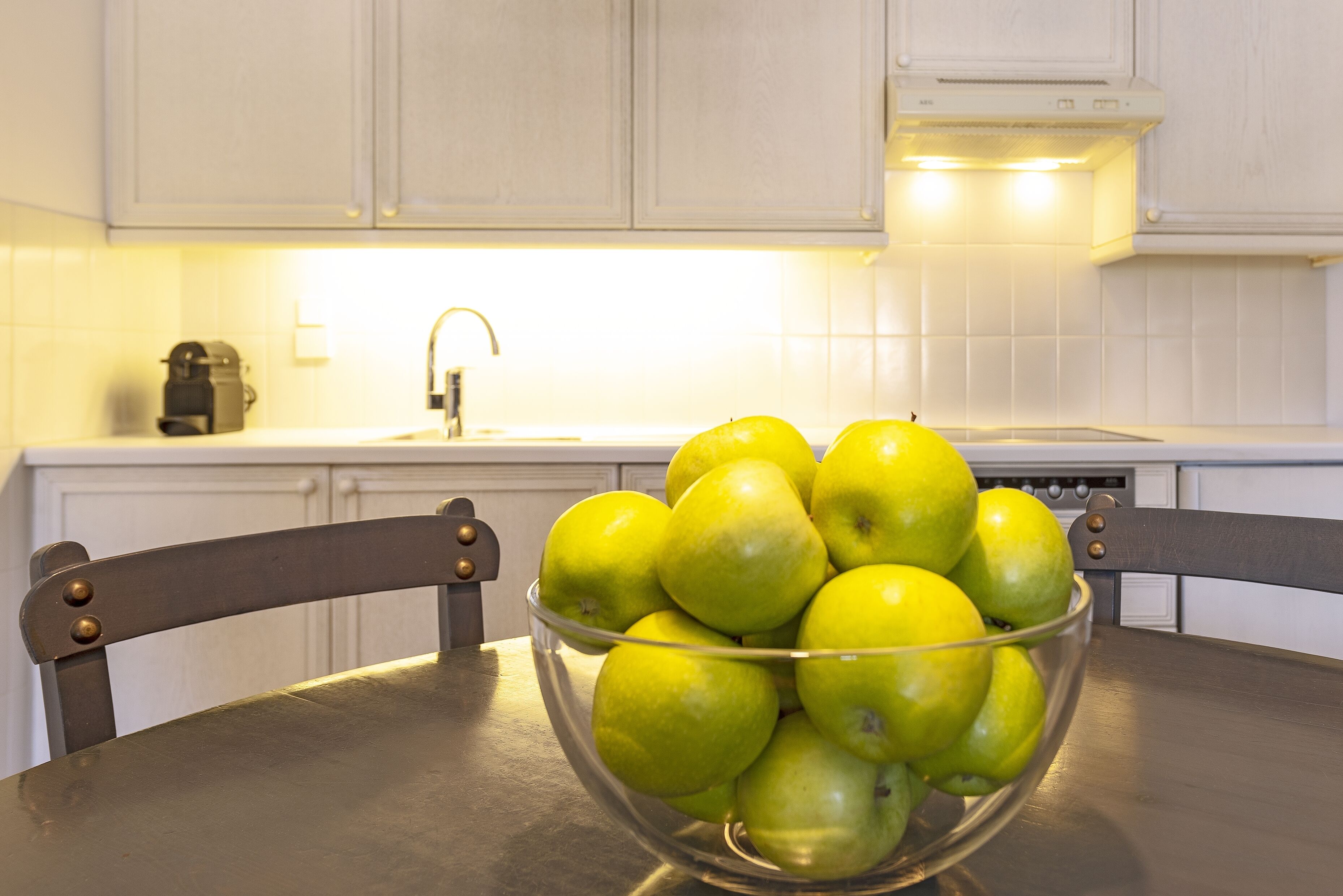 Een keuken met witte kastjes, een gootsteen en een schaal groene appels op een tafel.