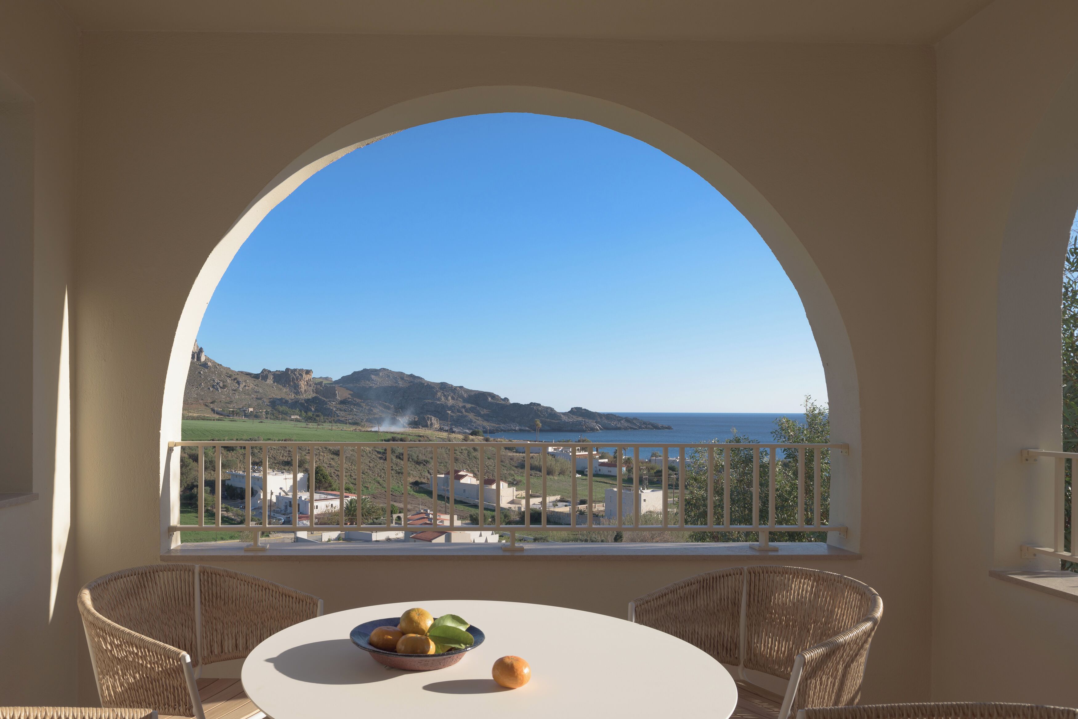 A balcony with wicker chairs, a round table with a bowl of fruit, and a view of the sea and hills.