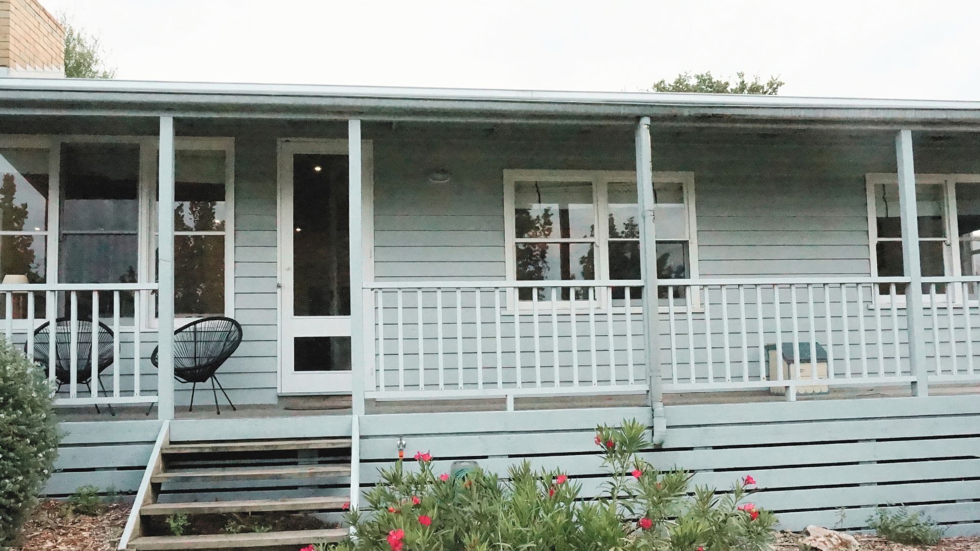 A light blue wooden house with a porch, white railings, and a small garden with red flowers.