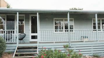 A light blue wooden house with a porch, white railings, and a small garden with red flowers.