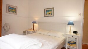 A bedroom with a bed, two bedside tables with lamps, a fan, and two framed pictures on the wall.