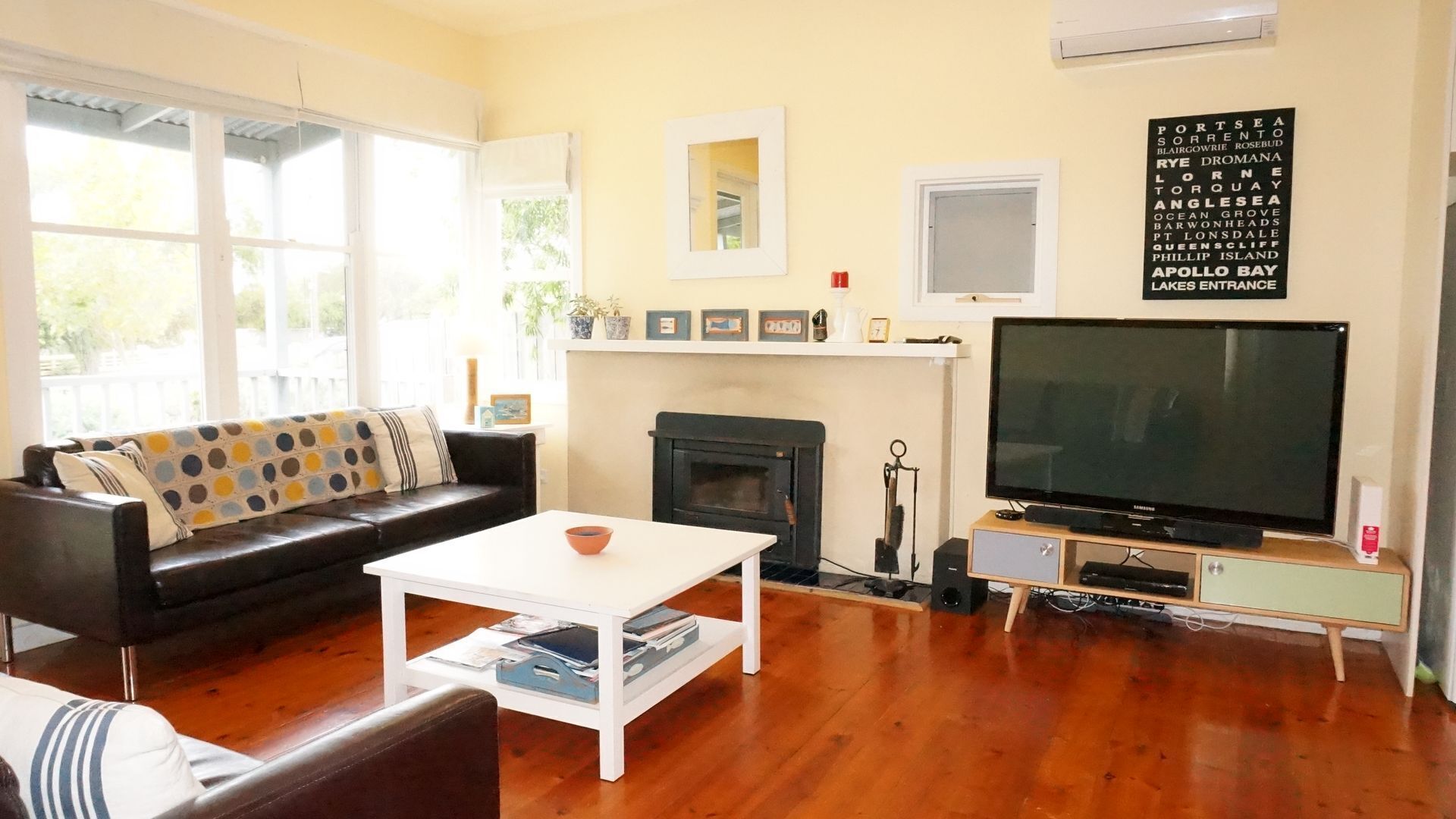 Una sala de estar con un sofá de cuero negro, una mesa de centro blanca, una chimenea, un televisor de pantalla plana grande y un espejo montado en la pared.