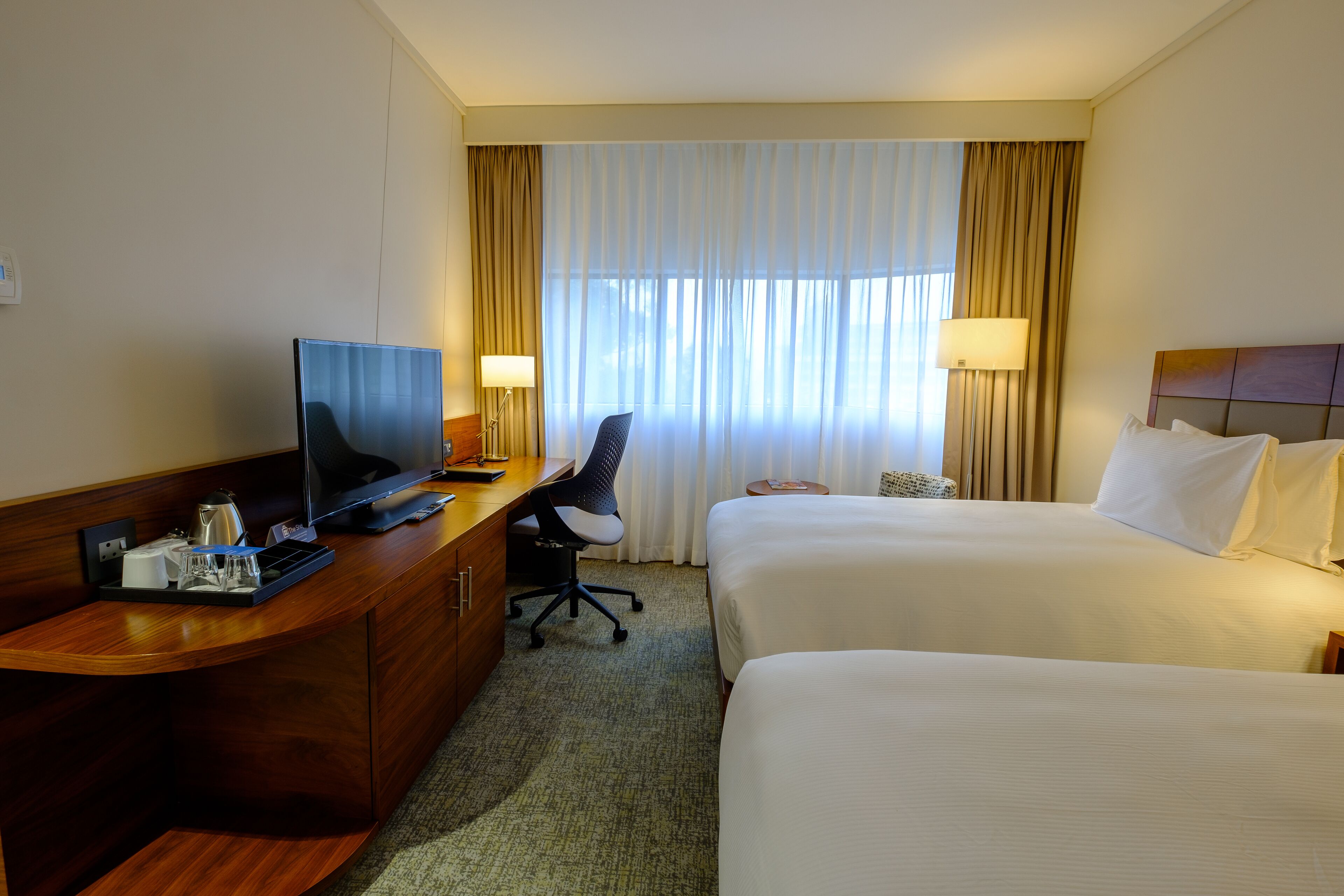 Ein Hotelzimmer mit Schreibtisch, zwei Betten, einem Fernseher und einem Fenster mit Vorhängen.