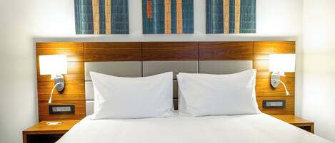 Une chambre d’hôtel avec une tête de lit en bois, deux oreillers blancs et une table de chevet avec une lampe.