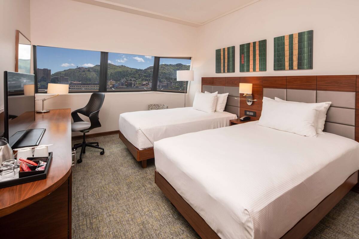 Quarto de hotel com duas camas, uma escrivaninha com computador, uma cadeira e vista para a cidade.
