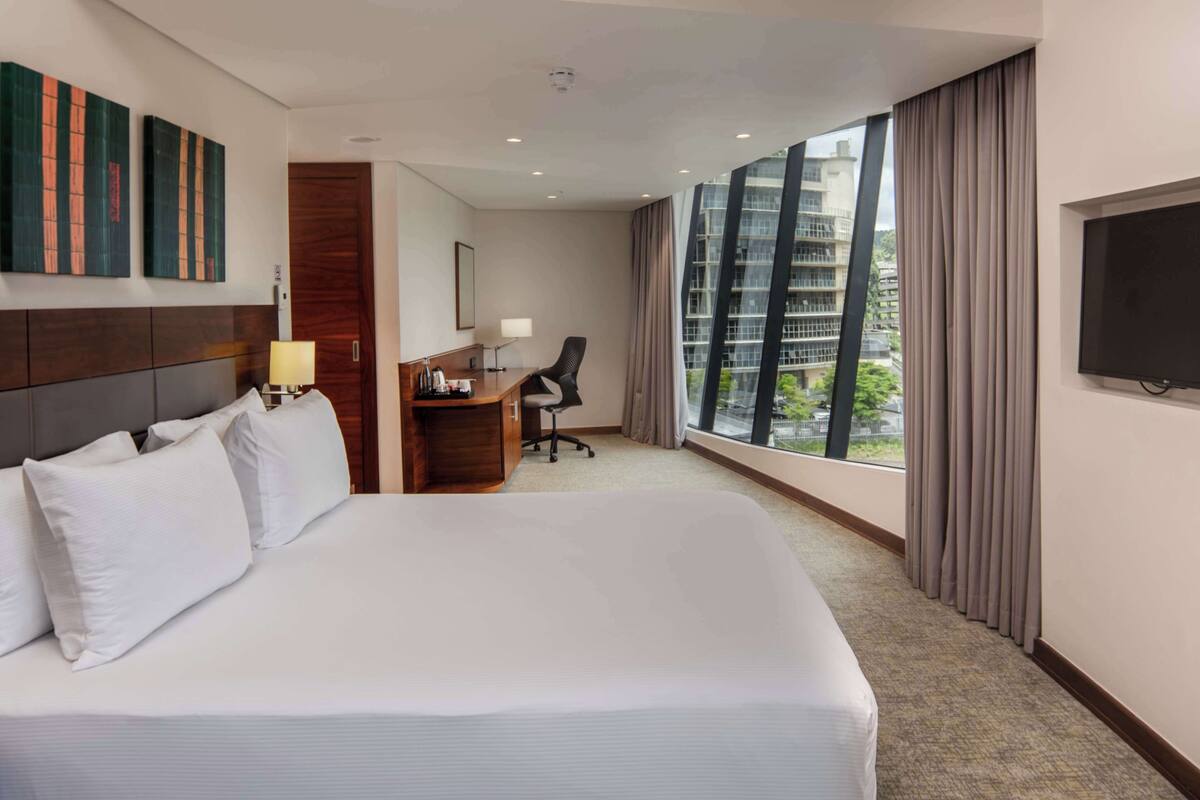 Quarto de hotel com uma cama grande, uma escrivaninha com cadeira, televisão e janela com cortinas.