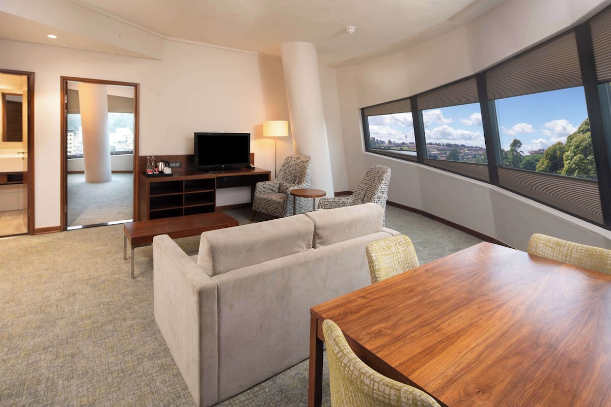 Quarto de hotel moderno com um sofá, uma mesa de jantar de madeira, uma TV e uma janela grande com vista para a cidade.