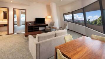 Una habitación de hotel moderna con un conjunto de sofás, una mesa de comedor de madera, un televisor y un ventanal con vistas a la ciudad.