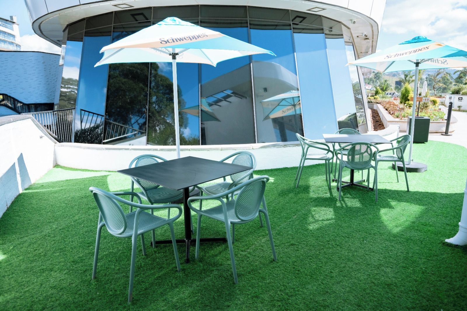 An outdoor seating area with artificial grass, metal chairs, and tables, and umbrellas with the brand 'Schweppes' visible.
