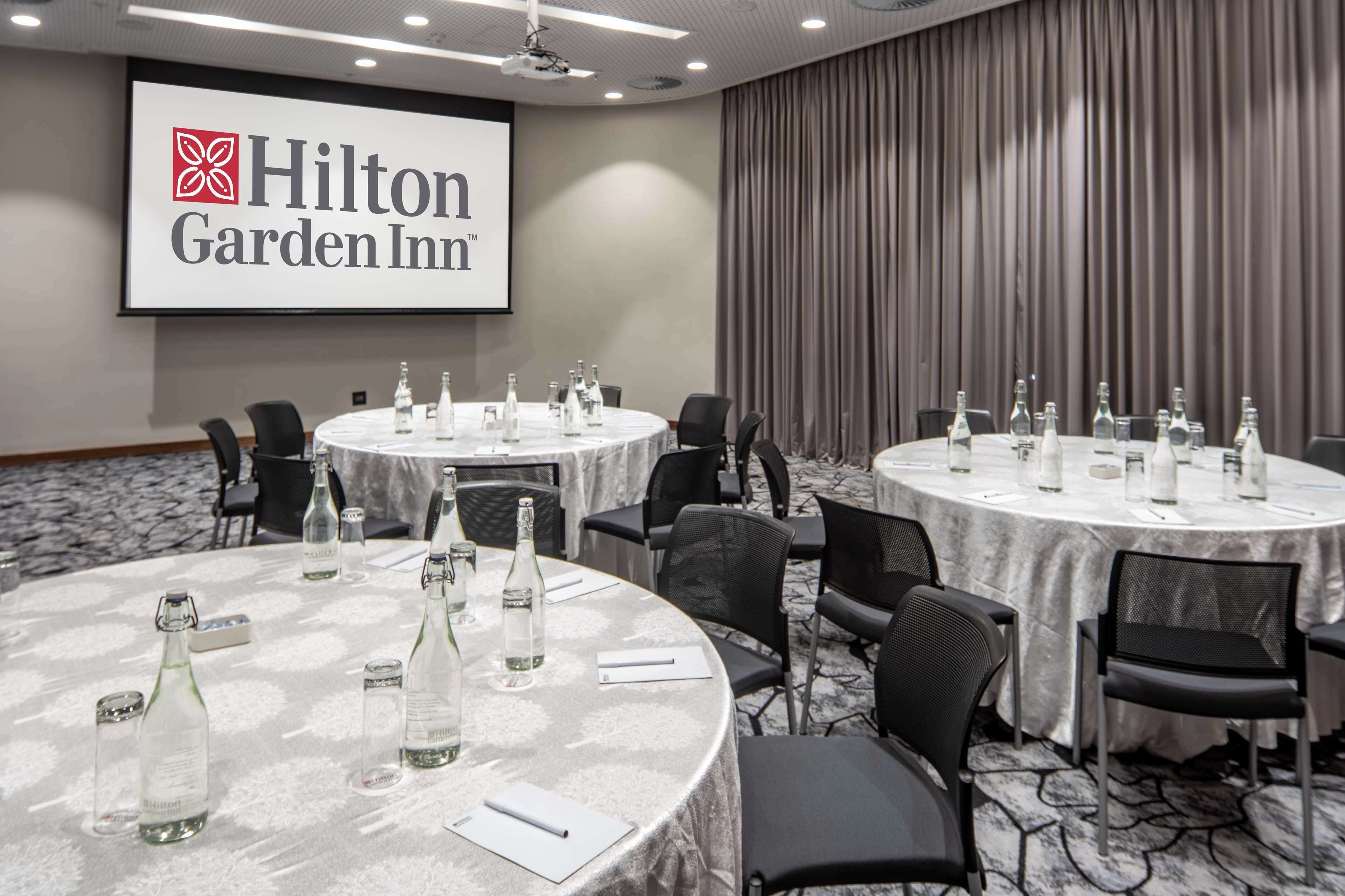 Een vergaderruimte met ronde tafels, stoelen en waterflessen, met het Hilton Garden Inn logo op de muur.
