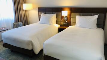 Hotellihuoneessa on kaksi sänkyä, joissa on valkoiset lakanat ja puinen pääty. Sänkyjen välissä on yöpöytä, jossa on lamppu, ja niiden lähellä on pieni pyöreä pöytä, jonka päällä on lehti.