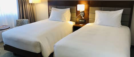 A hotel room with two beds, each with white linens and a wooden headboard. There is a nightstand with a lamp between the beds, and a small round table with a magazine on it.