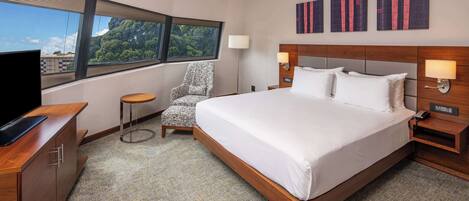 A hotel room with a large bed, a TV, a chair, and artwork on the wall.