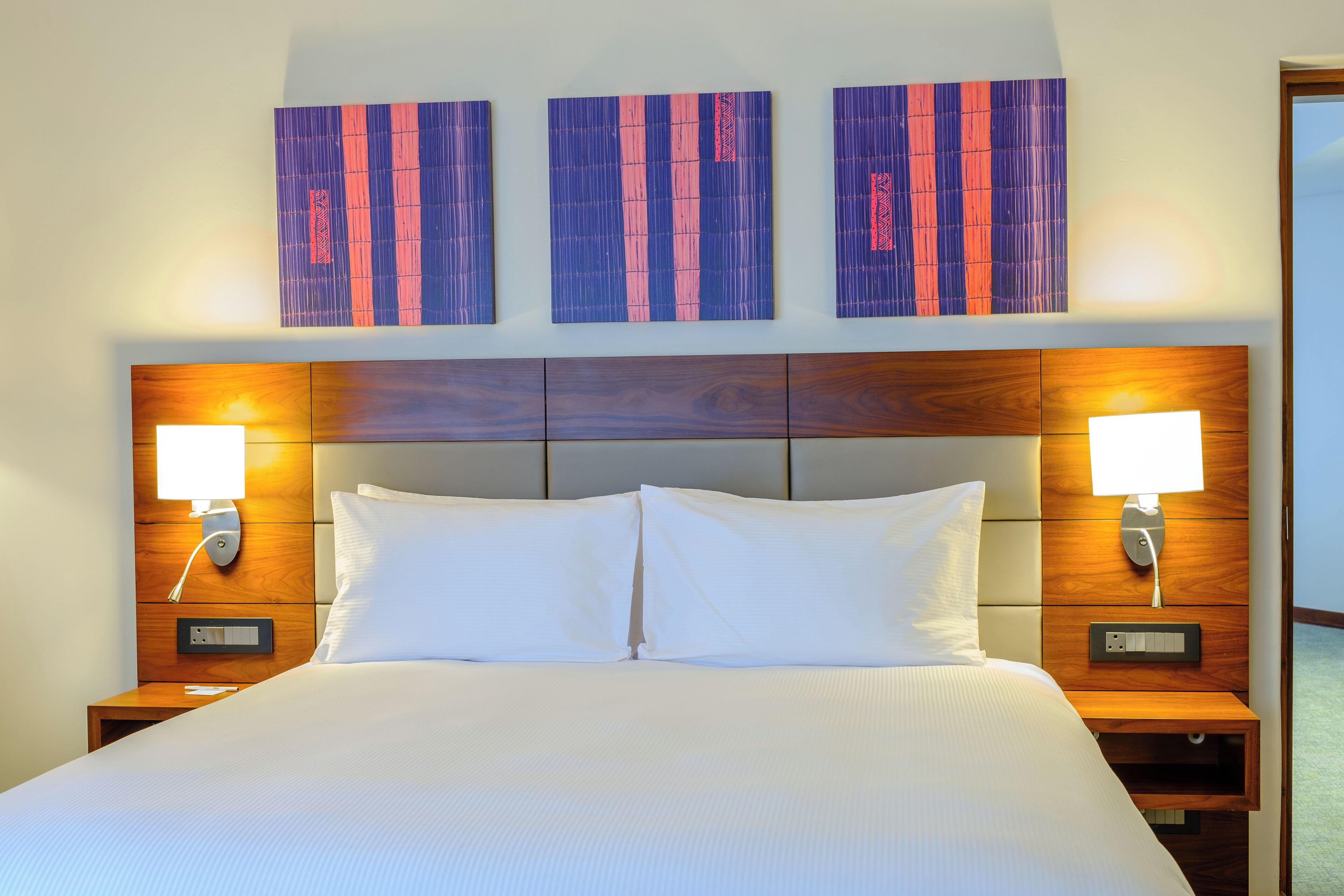 Habitación de hotel con una cama grande, dos mesitas de noche con lámparas y tres cuadros abstractos en la pared.