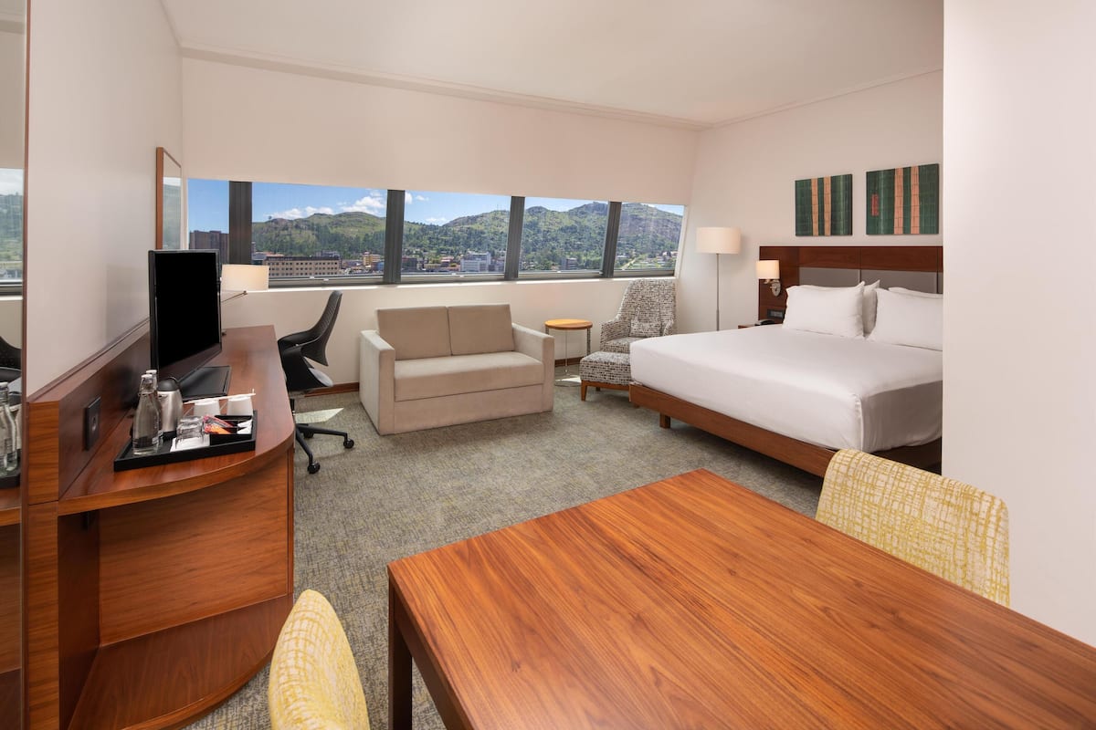 Quarto de hotel com cama, escrivaninha, cadeira, mesa pequena e vista para montanhas.