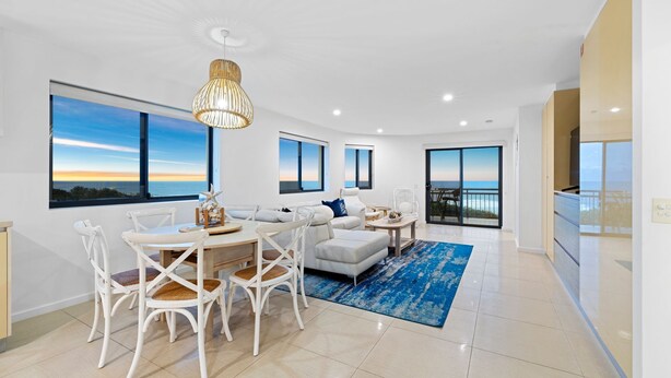 A modern living room with a dining area, large windows with ocean views, and a ceiling light fixture. - SHORELINE 9 PENTHOUSE WITH OCEAN VIEWS (HASTINGS POINT)