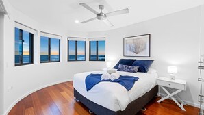 A modern bedroom with a large bed, multiple windows with ocean views, a ceiling fan, and a bedside table with a lamp. - SHORELINE 9 PENTHOUSE WITH OCEAN VIEWS (HASTINGS POINT)