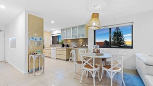 A modern kitchen with white cabinets, a dining area with white chairs, and a large window with a view of trees and a sunset. - SHORELINE 9 PENTHOUSE WITH OCEAN VIEWS (HASTINGS POINT)