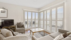 A modern living room with a flat-screen TV, a grey sofa, a wooden coffee table, and large windows with white blinds. - Resort on the Beach 4302 with resort Pool (KINGSCLIFF)