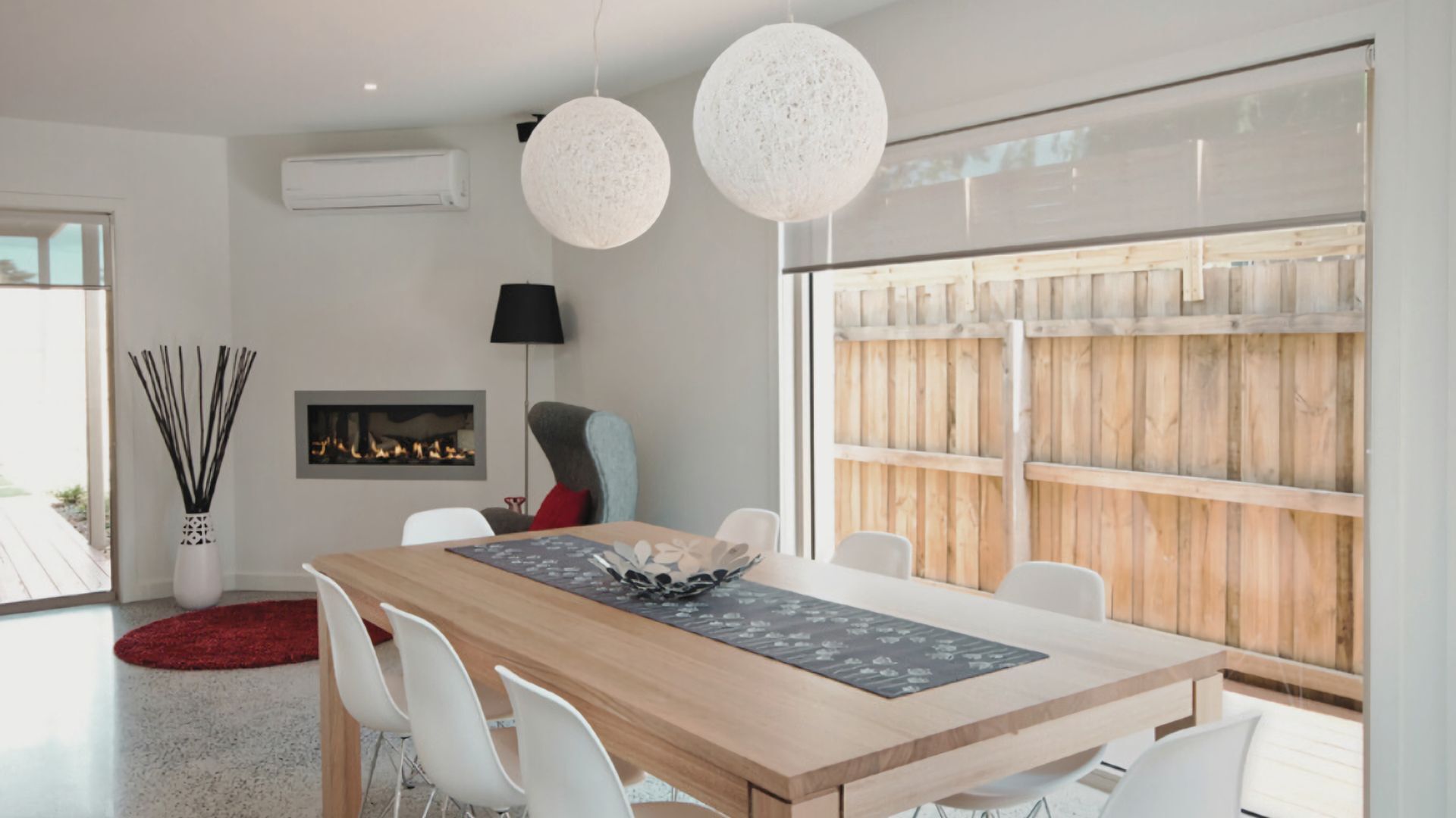 A modern dining area with a wooden table, white chairs, two spherical pendant lights, a fireplace, and a large window with a wooden fence view.