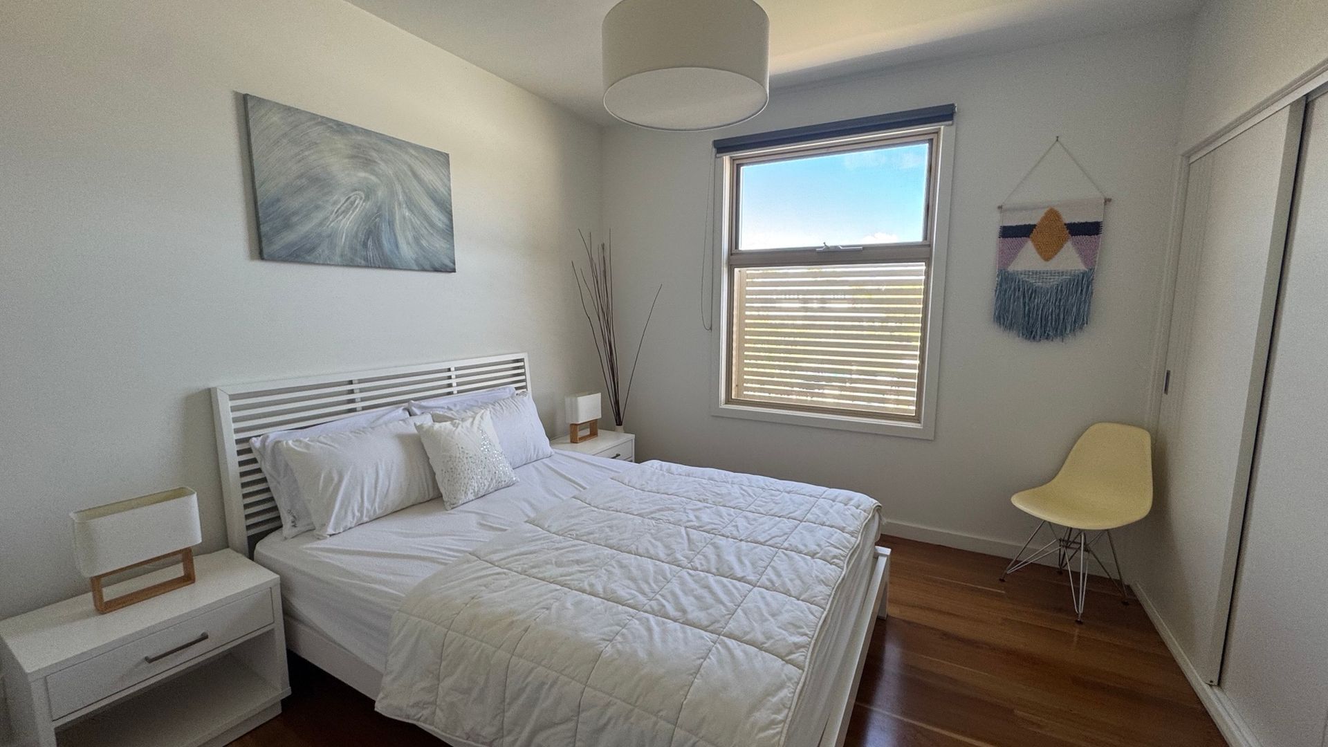 Une chambre à coucher comprenant un lit, une table de chevet, une lampe, une fenêtre et une œuvre d’art.