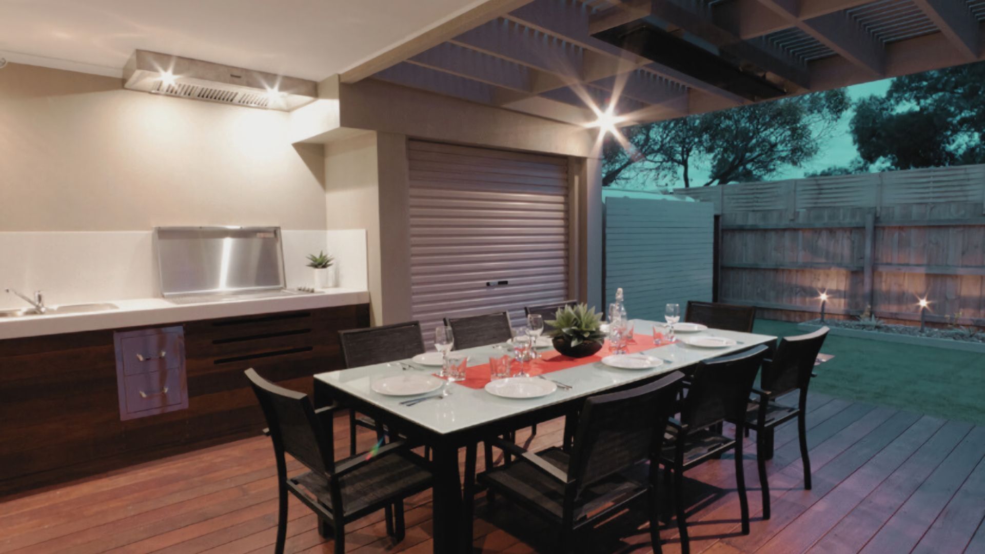 An outdoor dining area with a table set for six, surrounded by chairs, a built-in kitchen with a sink and stove, and a covered patio.