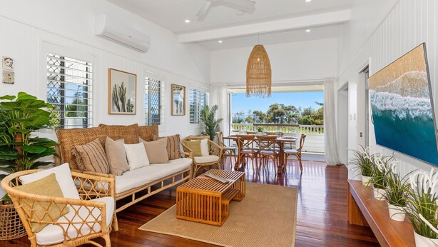A spacious living room with a sofa, chairs, a coffee table, and a dining area. There are large windows offering a view of the outdoors. - OCEANFRONT BEACH HOUSE ON MARINE PARADE (KINGSCLIFF)