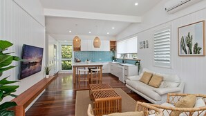 A modern kitchen with white cabinets, a blue backsplash, and wooden flooring. There is a dining area with a table and chairs, and a living room with a white sofa and a wicker chair. - OCEANFRONT BEACH HOUSE ON MARINE PARADE (KINGSCLIFF)