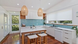 A modern kitchen with white cabinets, a blue backsplash, wooden stools, and two hanging wicker light fixtures. - OCEANFRONT BEACH HOUSE ON MARINE PARADE (KINGSCLIFF)