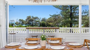 A dining table set for six with white plates and wine glasses, a vase with flowers, and a view of trees and a fence. - OCEANFRONT BEACH HOUSE ON MARINE PARADE (KINGSCLIFF)