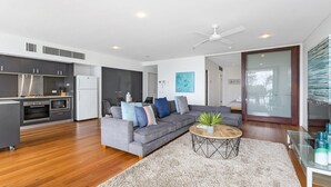 Una sala moderna con un sofá gris, una mesita de centro y una planta pequeña. Hay una zona de cocina con armarios oscuros y un refrigerador blanco.
