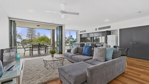 Un salón moderno con un sofá gris, una mesa de centro y un televisor sobre una mesa de vidrio. Hay un balcón con comedor y vistas al exterior.