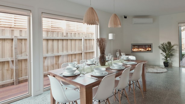 A dining area with a wooden table set for six, white chairs, pendant lights, a vase with branches, and a fireplace. - Taworri at Swan Bay in Queenscliff (Queenscliff)