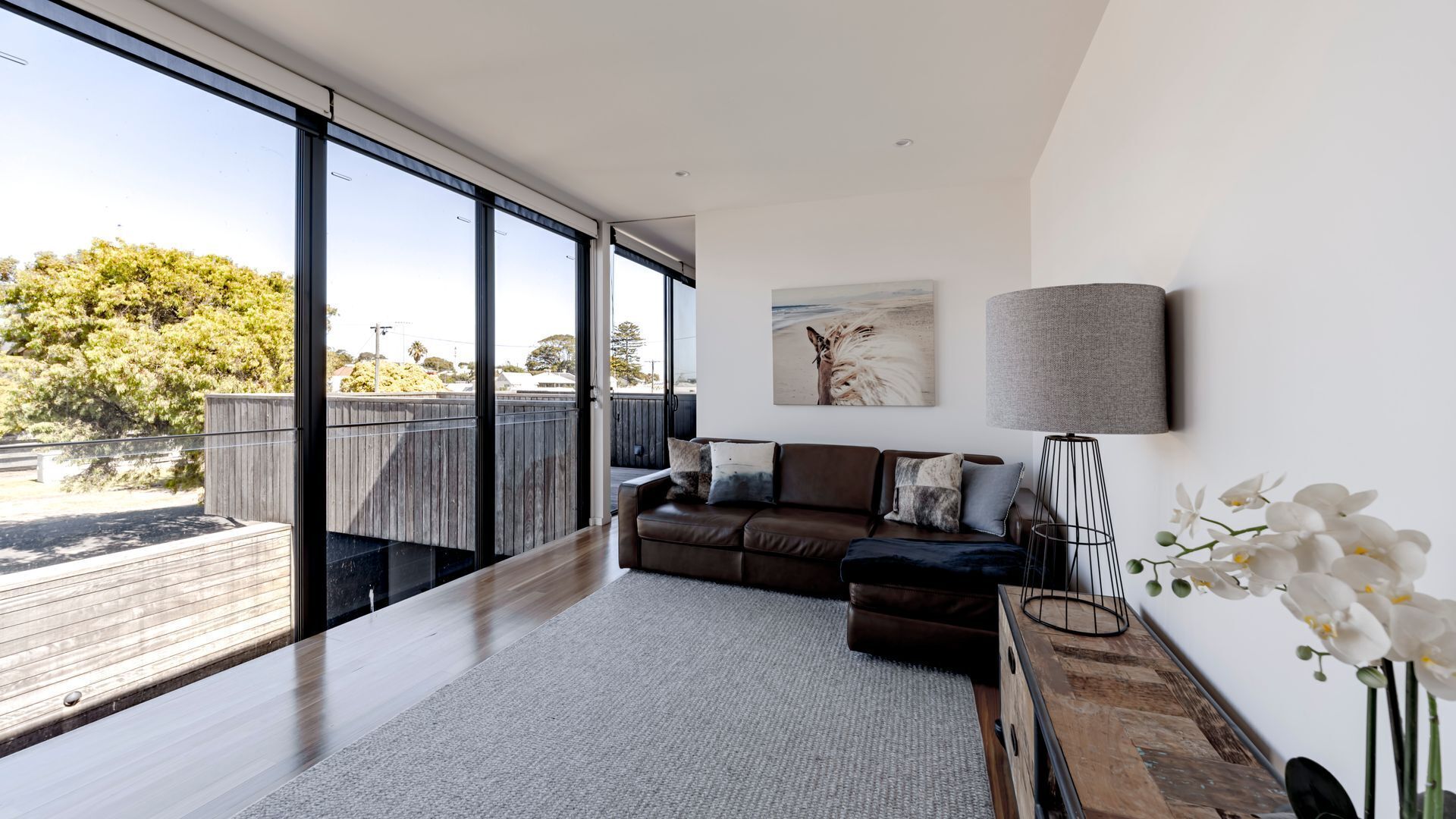 A modern living room with a large window, a brown leather sofa, a grey rug, and a minimalist lamp.