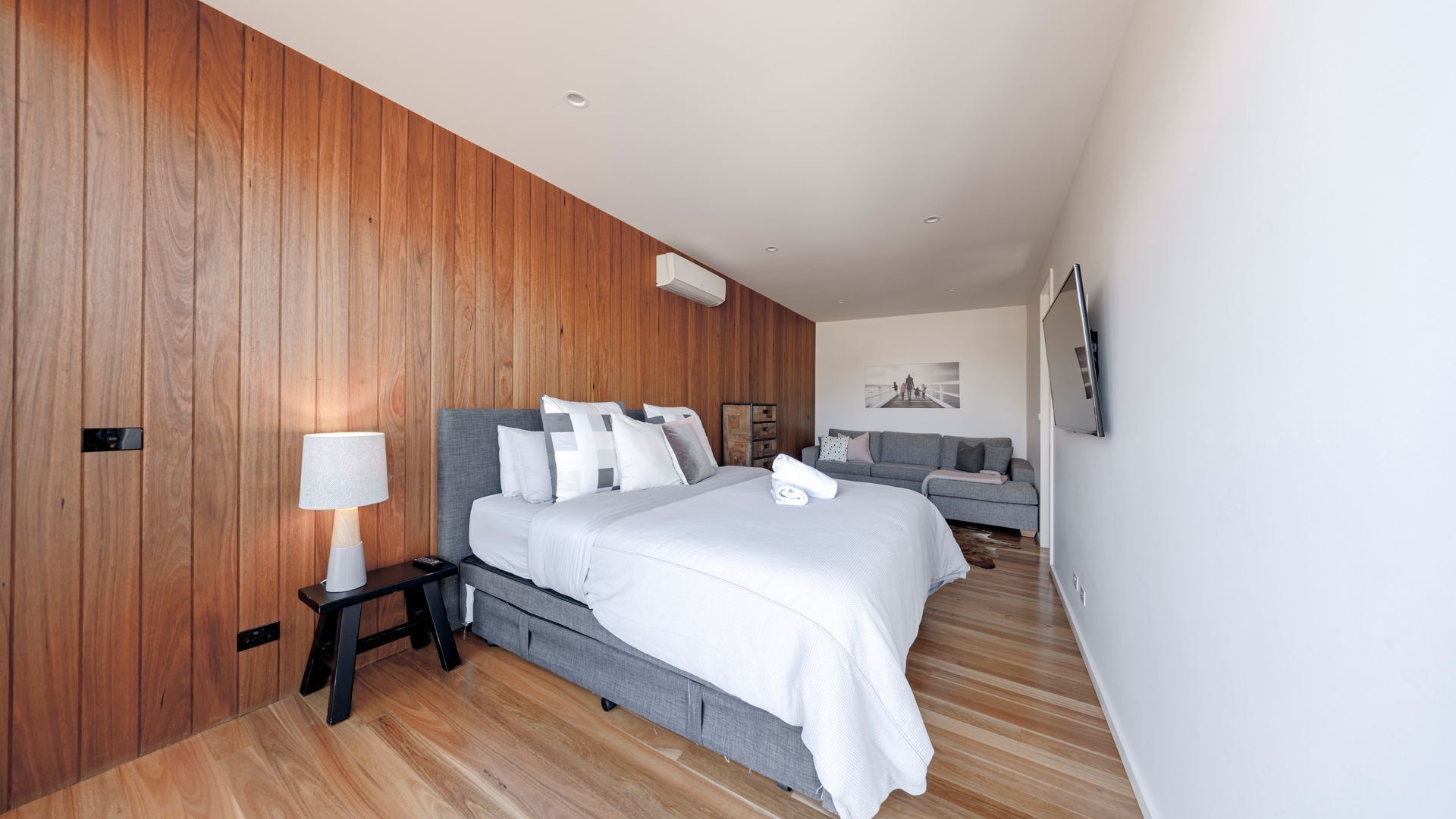 Ett modernt hotellrum med en väggpanel av trä, en säng med vita sängkläder, en grå soffa och en väggmonterad TV.