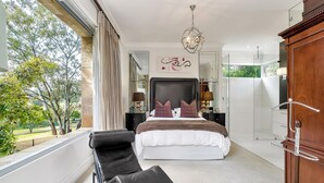 Une chambre avec un grand lit, un coin salon avec des fauteuils, une armoire en bois et une vue sur les arbres par la fenêtre.