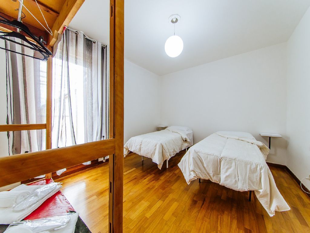 A room with two single beds, wooden flooring, a hanging light fixture, and a window with curtains.